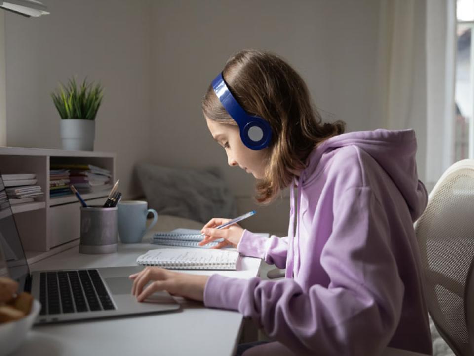 Teen girl school pupil wearing headphones studying online from home making notes.