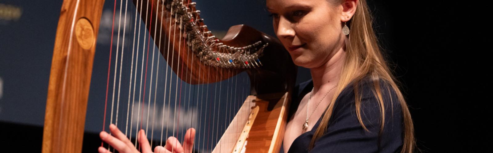 A woman with long blonde hair plays the Irish Harp in close up, her left hand is flat against the strings and her right hand is plucking